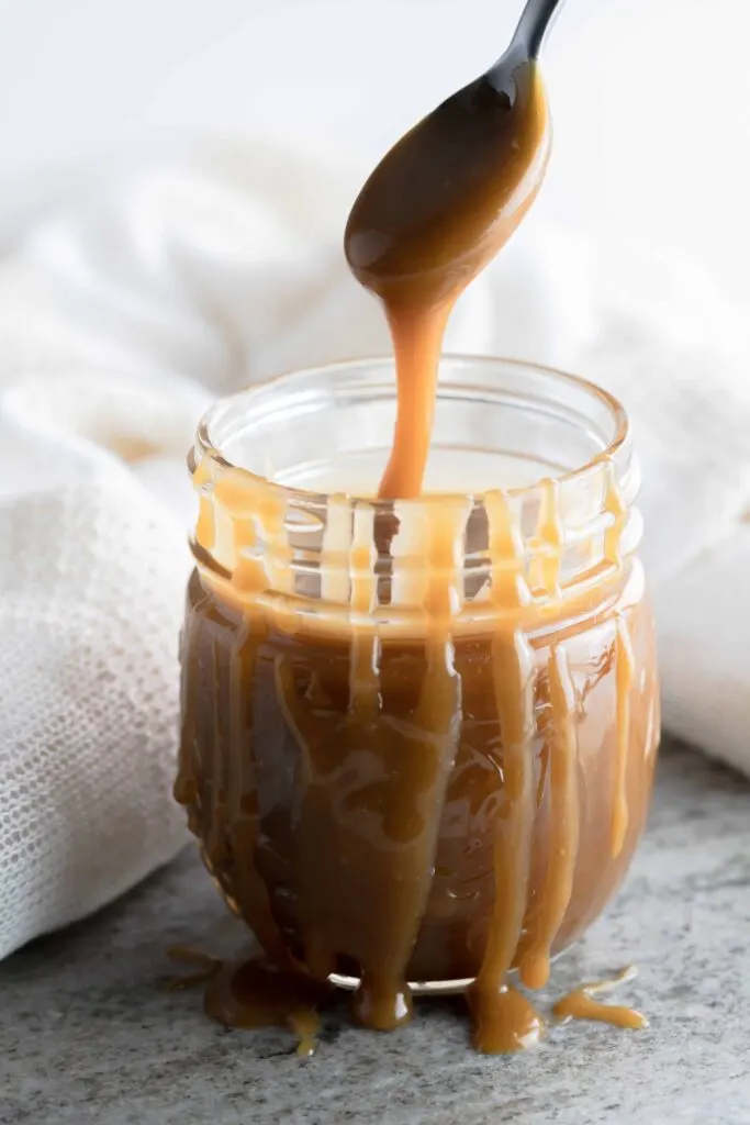 A jar of homemade butterscotch sauce with a spoon risen above the jar with sauce dripping back into the jar. 