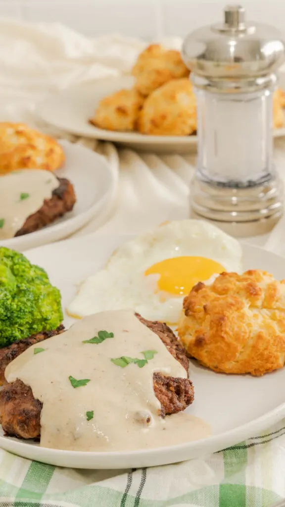 A drop biscuit on a plate with country fried steak covered with white gravy and garnished with parsley.