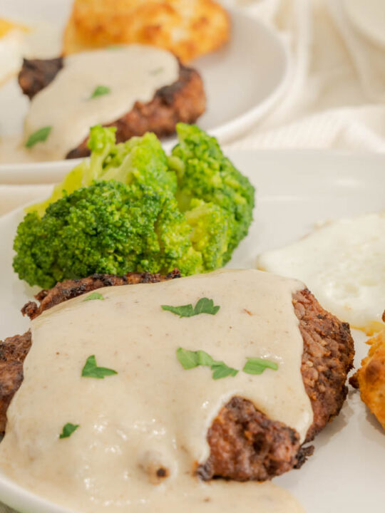 Country fried steak with gravy on a plate with broccoli in the background.