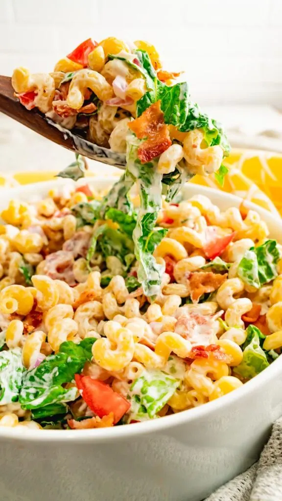 A spoon of blt pasta salad with dressing hovering above a bowl of pasta.