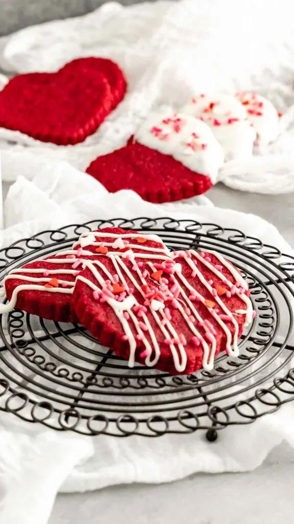 Two drizzled red velvet cookies on a round black decorative cooling rack with decorated cookies in the background.