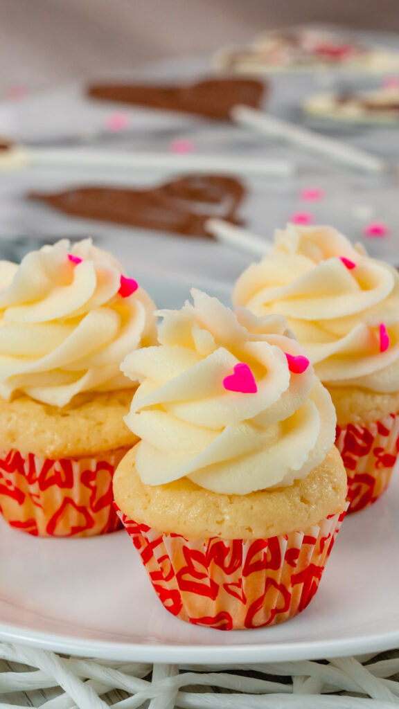 Three mini vanilla cupcakes on a white plate with a chocolate heart in the background.