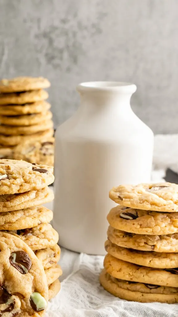 A white milk glass surrounded by stacks of chocolate chip cookies.