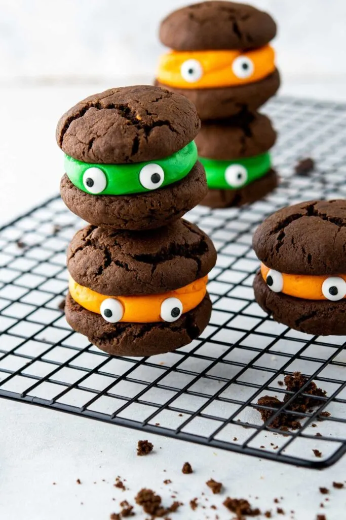 Easy Halloween Cookies stacked on a wire rack with green and orange buttercream filling and candy eyeballs.